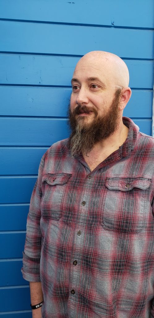 Nathan Kizerian lt. governor candidate of utah stands in front of blue buiolding he has a bald head and beard and plaid shirt