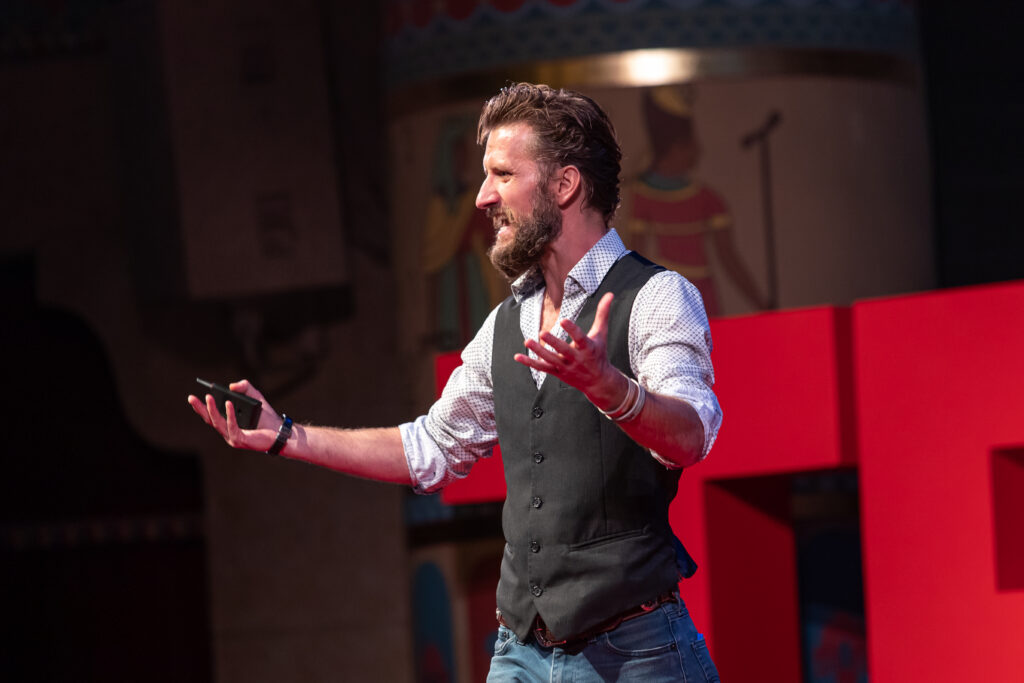 Zachary Moses speaking on the TEDxOgden stage, holding a clicker before red TEDx letters