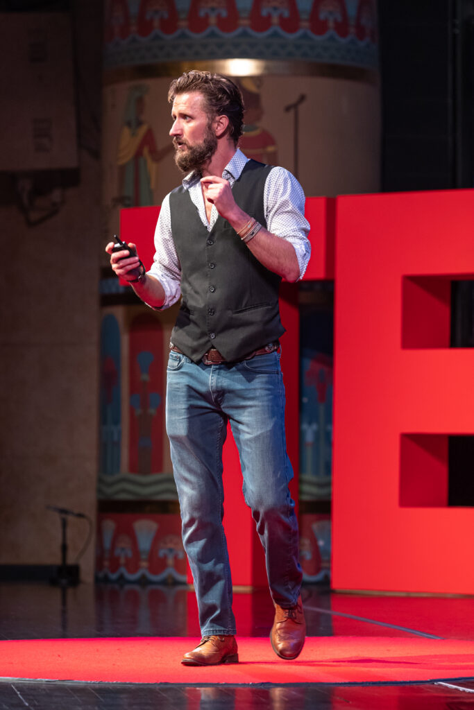 Zachary Moses speaking on stage at TEDxOgden in a white patterned shirt and dark vest